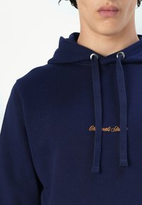 Navy blue hoodie made of smooth fabric, featuring a drawstring hood and silver eyelets. Includes subtle orange embroidered text.
