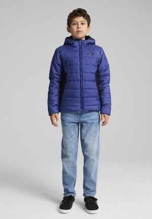 Niebieska kurtka puchowa z kapturem, z pikowanym wzorem, z przodu na zamek błyskawiczny i logo na piersi. W zestawie z jasnoniebieskimi dżinsami i czarnymi sneakersami.