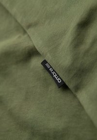 Khaki zelená bavlněná tkanina s jemnou texturou, s černou značkovou etiketou v rohu, která ukazuje minimalistický design.
