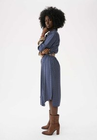 Dunkelblaues Kleid mit Punkten, langen Ärmeln und tailliertem Gürtel, kombiniert mit braunen Stiefeletten. Glattes Material mit fließender Silhouette.