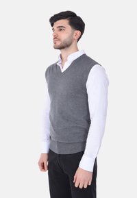 Gilet in maglia grigia con scollo a V e orlo a coste, indossato sopra una camicia bianca a maniche lunghe. Texture liscia, forma aderente.