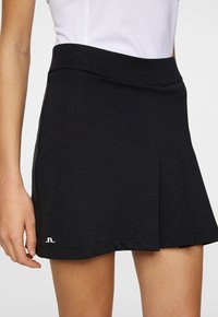 Svart tenniskjol med en utställd design, tillverkad av lätt material. Har en bred midja och en liten logotyp på fållen.