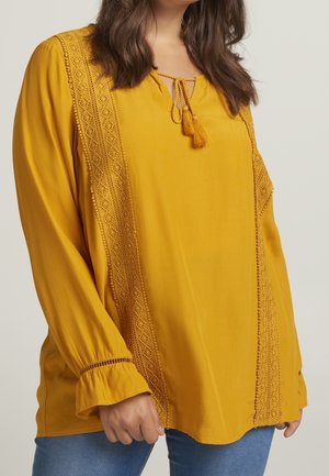 Femme portant un chemisier jaune moutarde avec des détails en dentelle et un lien à pompons, associé à un jean bleu, visible des épaules aux hanches.