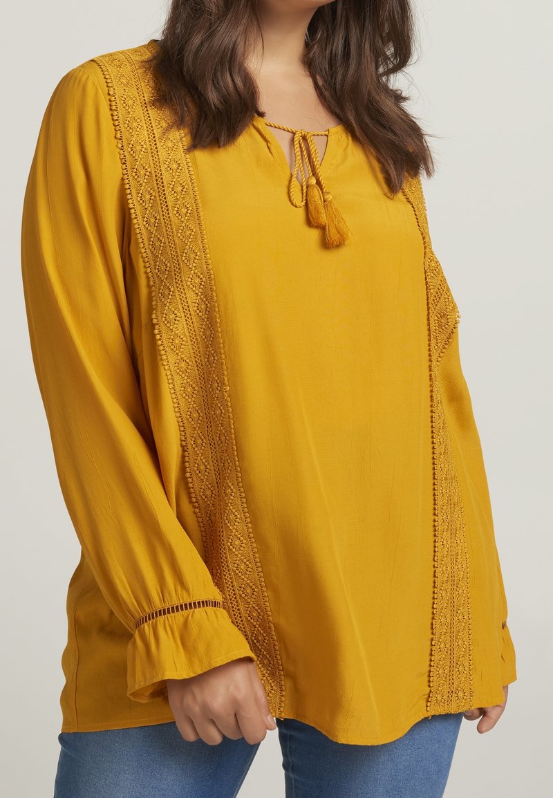 Femme portant un chemisier jaune moutarde avec des détails en dentelle et un lien à pompons, associé à un jean bleu, visible des épaules aux hanches.