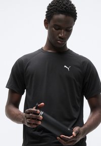 Μαύρο αθλητικό κοντομάνικο μπλουζάκι με λευκό λογότυπο Puma στο στήθος· το μοντέλο κρατά μπουκάλι νερού ματ μαύρο.