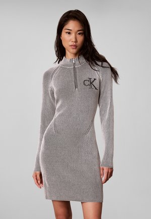 Frau trägt ein langärmliges, knielanges, schwarz-weiß gestreiftes Kleid mit einem Reißverschluss vorne und dem "cK"-Logo auf der Brust.