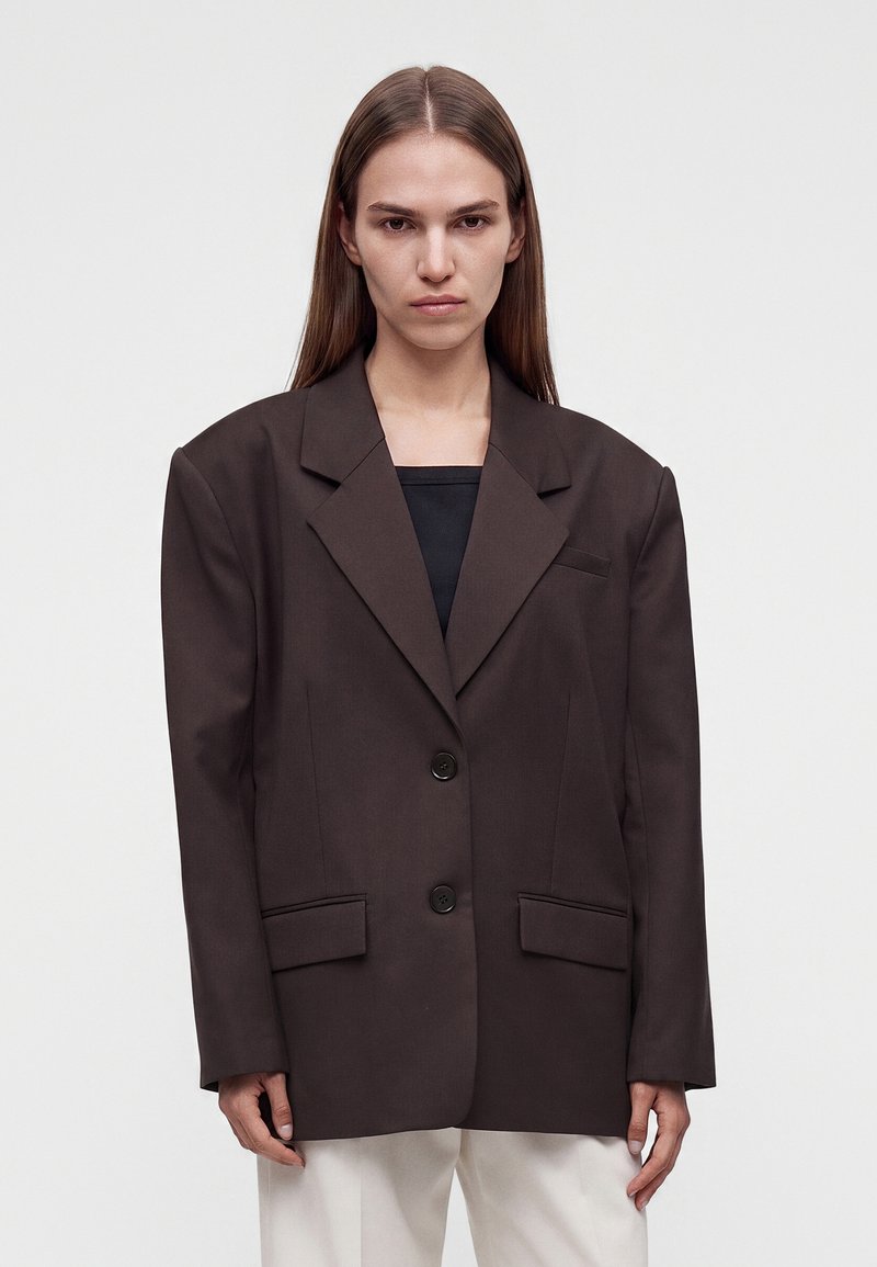 Femme aux longs cheveux raides portant un blazer marron foncé oversize sur un haut noir et un pantalon blanc, debout devant un fond uni.