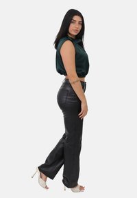 Kobieta z długimi ciemnymi włosami, ubrana w bezrękawnik w ciemnozielonym kolorze, czarne skórzane spodnie, białe szpilki i tatuaż na ramieniu, pozująca z boku.