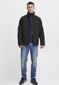 Sort jakke med høj krave, lynlåslukning og subtilt logo, parret med en blå sweater og lyseblå jeans. Bæret med mørke sneakers.