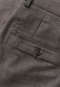 Gros plan sur un tissu à carreaux marron montrant une poche arrière boutonnée d'un pantalon sur mesure avec des détails cousus et une passants de ceinture.