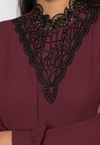 Bordeauxblus med intrikata svarta spetsbroderier runt halslinningen och mjukt, texturerat tyg. Figursydda ärmar med milda volanger.