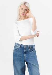Hvit, tettsittende langermet topp med båthals, kombinert med høytlivede blå denimjeans med en avslappet passform og falmet tekstur.