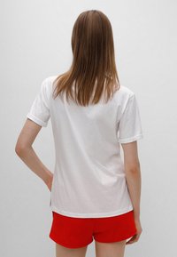 Hvid bomulds T-shirt med oprullede ærmer, set fra ryggen. Kombinationen med røde shorts fremhæver et simpelt, afslappet design.