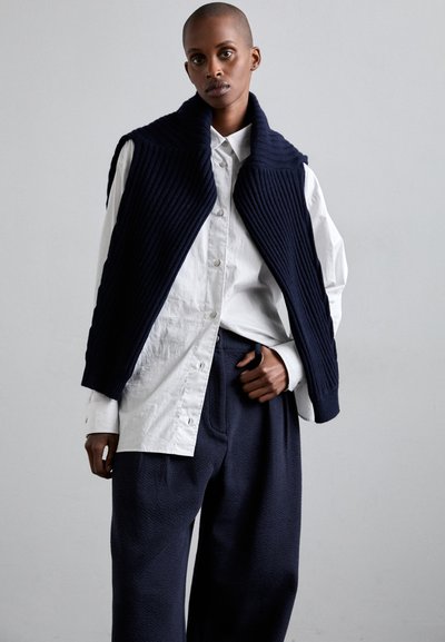 Gilet a coste blu navy con colletto alto sopra una camicia bianca con bottoni, abbinato a pantaloni scuri blu navy a trama. Design semplice e moderno.