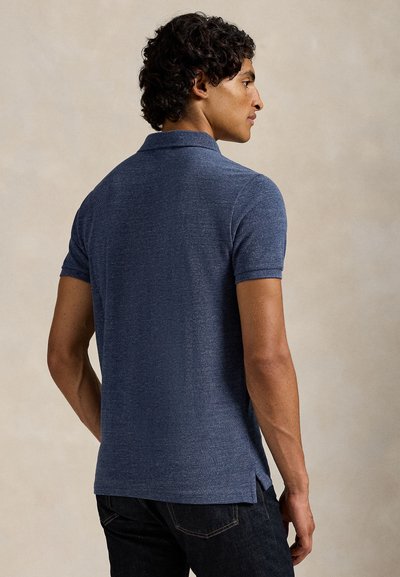 Homme aux cheveux courts et bouclés portant un polo bleu à manches courtes et un jean foncé, debout, tourné de dos sur un fond uni.
