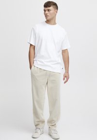 T-shirt blanc à manches courtes avec une coupe décontractée, associée à un pantalon beige en velours côtelé. Le mannequin porte des baskets blanches, les mains dans les poches.