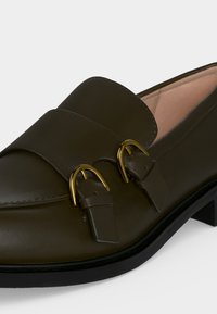 Σκούρο πράσινο δερμάτινο loafer με δύο χρυσές αγκράφες, χαμηλή μαύρη φτέρνα και ανοιχτό μπεζ εσωτερική επένδυση.