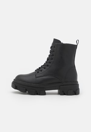 Botte de combat en cuir noir avec semelle crantée épaisse, laçage latéral et languette à l'arrière, vue de profil sur fond blanc.
