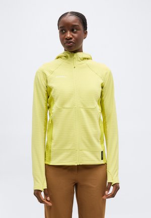 Žena v žltej mikine so zipsom a kapucňou, s bočnými vreckami a hnedými nohavicami, stojaca pred jednoduchým bielym pozadím.