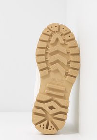 Semelle de botte en caoutchouc beige avec un motif de bande adhérente profond et le logo Palladium centré sur un fond blanc.