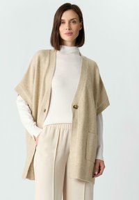 Beżowy sweter z dzianiny z krótkimi rękawami, dwoma przednimi kieszeniami, zapinany na pojedynczy guzik oraz z prążkowanymi akcentami wokół dolnej części i rękawów.