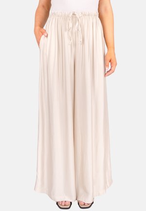 Femme portant un pantalon large et ample couleur crème avec une taille élastique et un cordon de serrage, une main dans la poche, associé à des chaussures noires à bout ouvert.