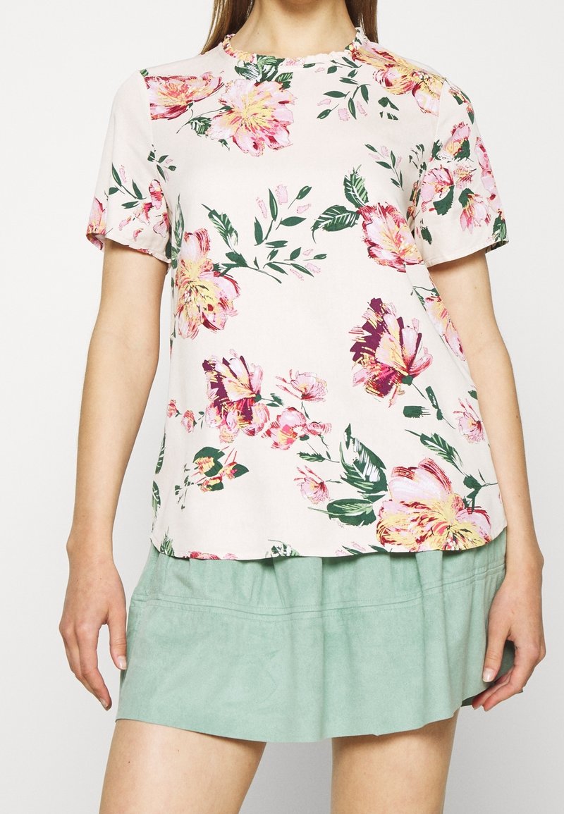 Femme portant un chemisier blanc à manches courtes avec un imprimé floral rose et jaune, associé à une jupe vert clair longueur genou.
