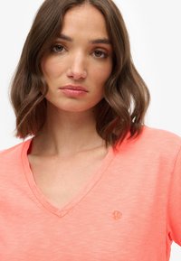 Koraal kleurige v-hals t-shirt gemaakt van zachte, lichte stof. Kenmerkt zich door een geborduurd logo aan de linkerzijde. Overgangsstructuur met een ontspannen pasvorm.