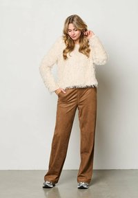 Romantische, luchtige trui met een cropped ontwerp, gecombineerd met brede, suède achtige broek in bruin en sneakers. Model poseert met één hand in de zak.