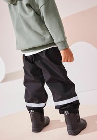 Pantalones impermeables negros con tiras reflectantes blancas, combinados con botas de goma negras. La parte superior es un hoodie de forro polar verde.