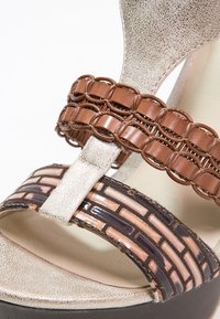 Gros plan d'une sandale beige et marron avec une bride en cuir tressé et une bride avant texturée à motifs géométriques au-dessus de l'orteil ouvert.