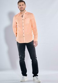 Hellorange Hemd mit durchgehendem Knopfverschluss und einer einzelnen Brusttasche, schwarzen Knöpfen, kombiniert mit dunklen Jeans und weißen Sneakers. Glatte Textur, taillierte Passform.