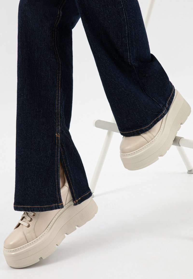 Tmavě modré džíny s širokými nohavicemi a postranním rozparkem, nošené s béžovými robustními botami na platformě se šněrováním, na bílém pozadí.