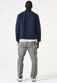 Marineblau gesteppte Jacke mit einem gerippten Kragen, kombiniert mit hellgrauen Hosen. Das Outfit besteht aus einem schlichten weißen Hemd darunter.