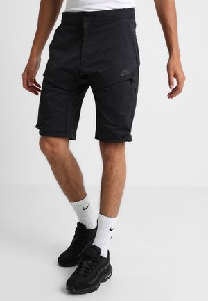 Sorte atletiske shorts med en glat tekstur, der har en subtil sidelomme med lynlås og et lille logo på låret. Båret med hvide sokker og sneakers.