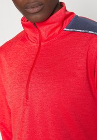 Röd halvzip-tröja tillverkad av ett texturerat tyg, med en mörkblå accent på axeln och diskret varumärkesprofilering på kanten.