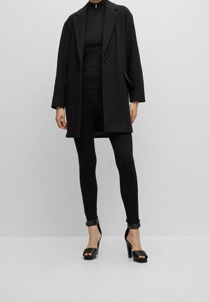 Persoon draagt een zwart op maat gemaakte jas, een zwarte top met rits, zwarte legging en zwarte sandalen met hoge hakken tegen een effen grijze achtergrond.
