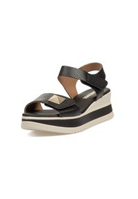 Sandalias de cuña de cuero negro con tiras cruzadas, un acento en forma de pirámide dorada y una suela blanca gruesa con una superficie texturizada.