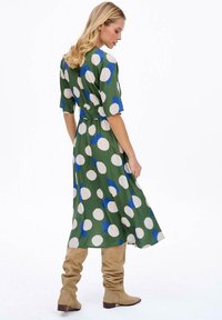 Zelené šaty s velkými bílými a modrými puntíky, krátké rukávy, stáhnutý pas, volný střih. Nosič má na sobě béžové kozačky po kolena.
