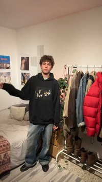 Jeune homme en sweat à capuche noir et jean large se tient dans la chambre près du lit et du portant à vêtements avec des vestes et des bottes, avec une plante d'intérieur en arrière-plan.