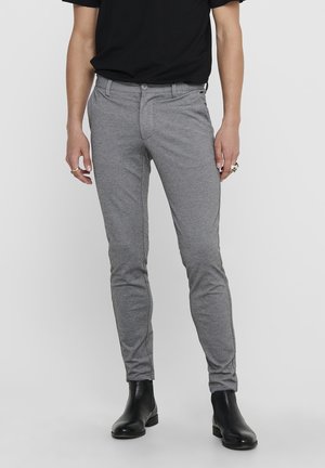 Man draagt slank gesneden grijze broek, zwart T-shirt en zwarte leren enkellaarzen, staand tegen een effen lichte achtergrond.