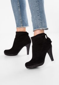 Bottes de cheville en daim noir avec un design effilé, un gland décoratif et un talon noir pailleté, associées à un jean bleu clair effiloché.