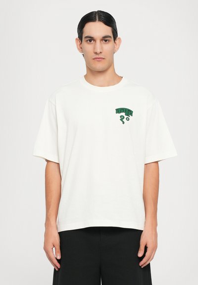 T-shirt branca de algodão com mangas curtas, apresentando um desenho gráfico verde na área do peito esquerdo. Corte reto na bainha.