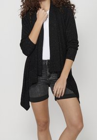 Svart öppen cardigan med metalliska prickar, långa ärmar och en avslappnad passform, i kombination med grå denimshorts. Texturen är ribbad och mjuk.