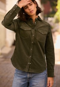 Camicia in corduroy verde oliva con chiusura a bottoni, tasche sul petto e bottoni a pressione argentati; maniche lunghe e dettaglio dell'orlo curvo.