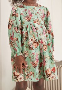 Květinové šaty z látky v mátově zelené barvě, s dlouhými rukávy, zapínáním na knoflíky a převázaným pasem. Po celém modelu se nacházejí akcenty růžových a hnědých květin.