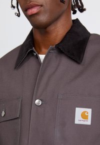 Grå jakke med en kordfløjskrave, sølvknapper og forlommer. Har et Carhartt-logo patch i orange og hvid.