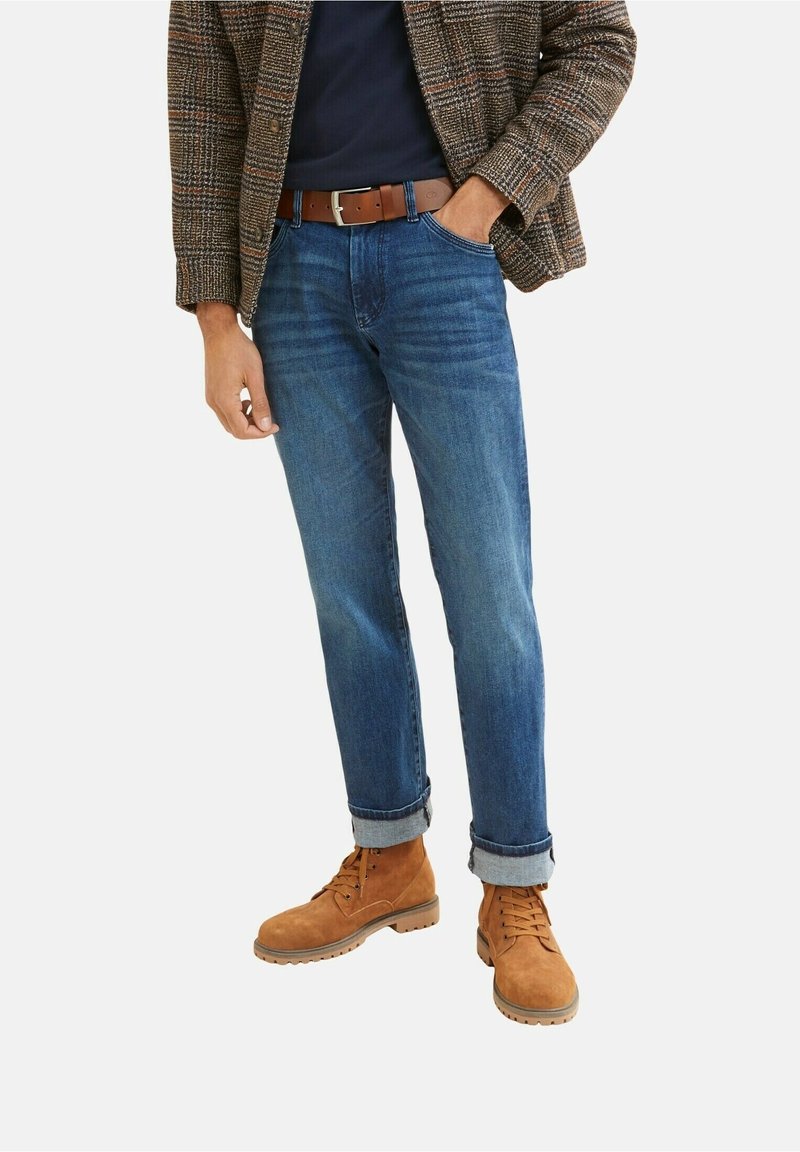 Homme portant un jean bleu retroussé, une ceinture en cuir marron, un blazer à carreaux, une chemise foncée et des bottes en daim beige lacées, debout avec une main dans la poche.