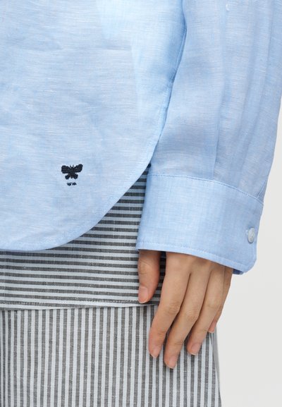 Poignet de chemise bleu clair à manches longues avec bouton, abeille noire brodée près de l’ourlet, main reposant à côté d’un tissu rayé verticalement gris et blanc.