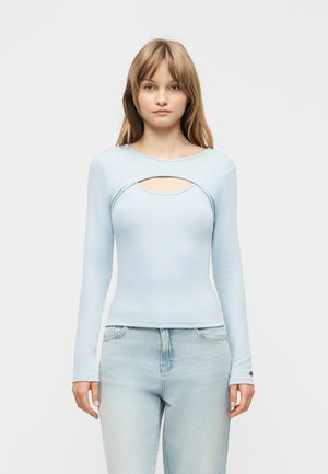 Mlada ženska, oblečena v svetlomodro majico z dolgimi rokavi in izrezom na prsih ter svetlo opranimi hlačami iz jeansa, stoji pred enobarvnim ozadjem.